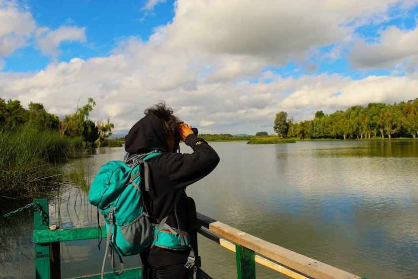 A person with a backpack standing on a green bridge over a calm lake, surrounded by trees and a partly cloudy sky. 1