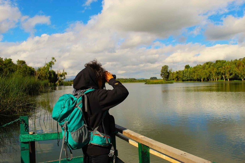 A person with a backpack standing on a green bridge over a calm lake, surrounded by trees and a partly cloudy sky. 2