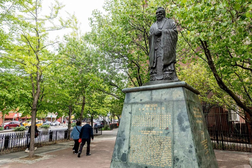 A statue of Confucius on a pedestal with inscriptions, surrounded by trees and a sidewalk with pedestrians. 5