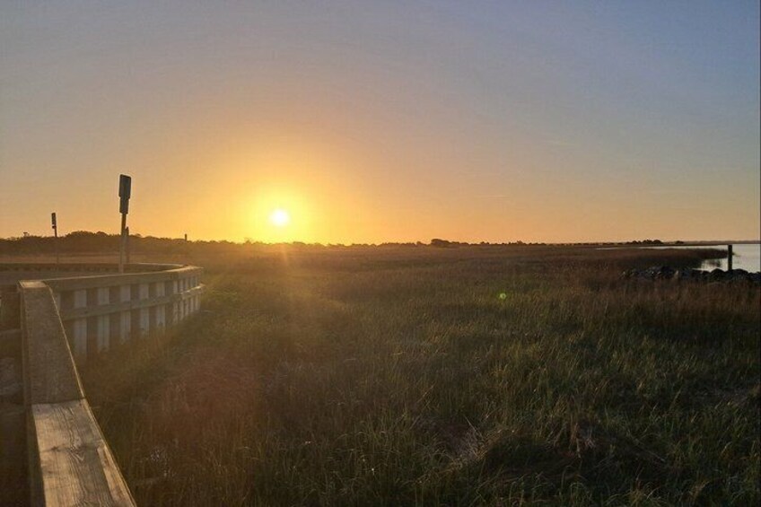 A sunset over a grassy field with a road barrier and a distant water body. 2