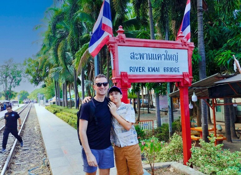A man and a child standing in front of a sign that reads 'River Kwai Bridge' with Thai text, palm trees, and a railway track in the background. 2
