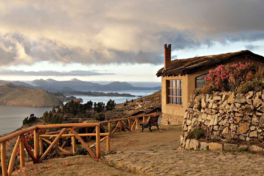 A stone house with a wooden fence and bench, overlooking a lake and mountains. 3