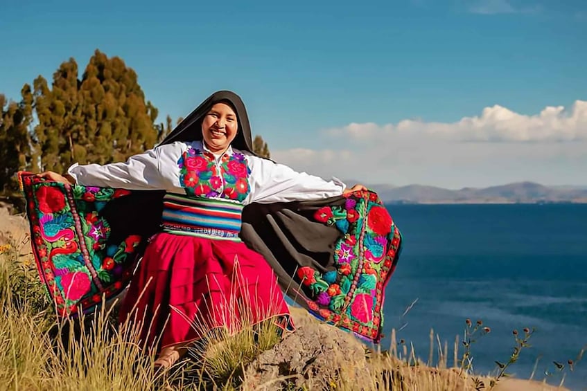 A person in traditional attire sitting on a rock by a lake with mountains in the background. 1