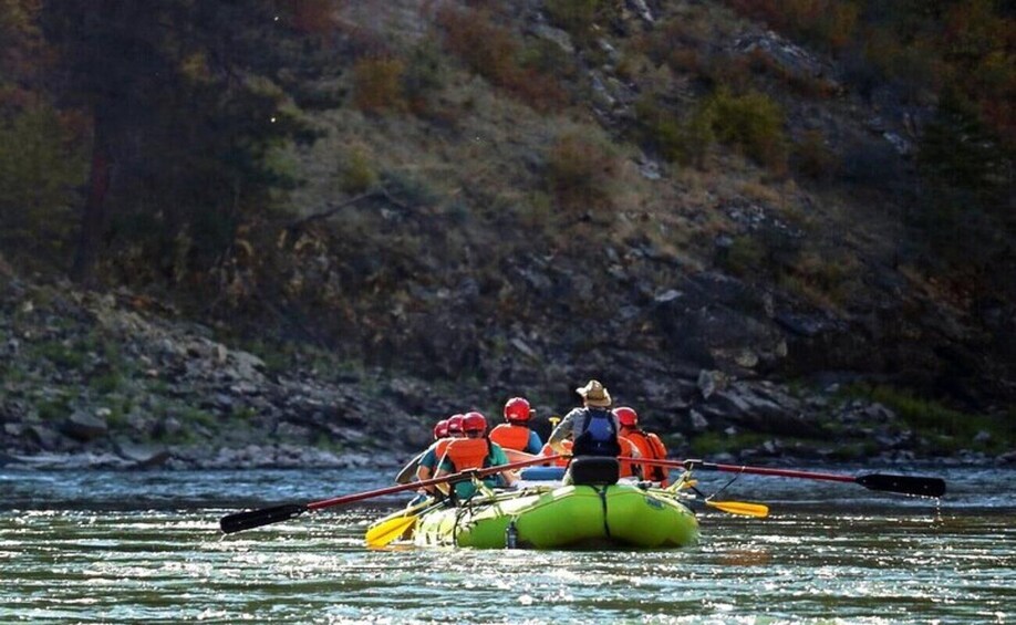 A group of people in a green raft on a river, paddling with oars. 3