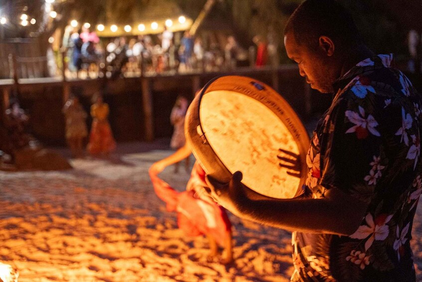 A person holding a round, flat object with a textured surface, possibly a musical instrument, in a dimly lit outdoor setting with a crowd in the background. 5