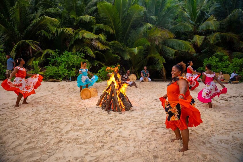 A group of people dancing around a fire on a sandy beach with palm trees in the background. 1