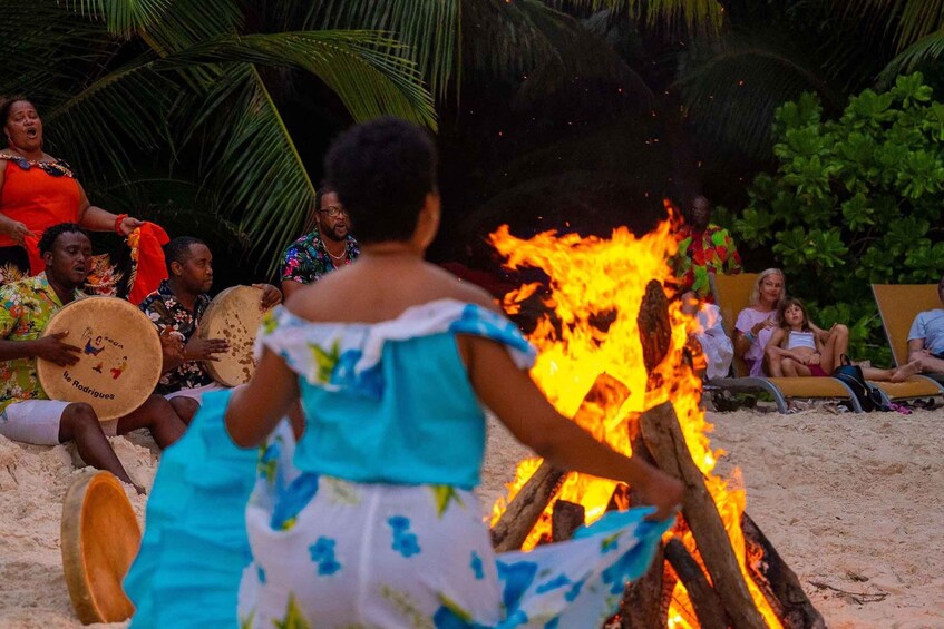 A beach scene with a fire, people playing drums, and palm trees. 4