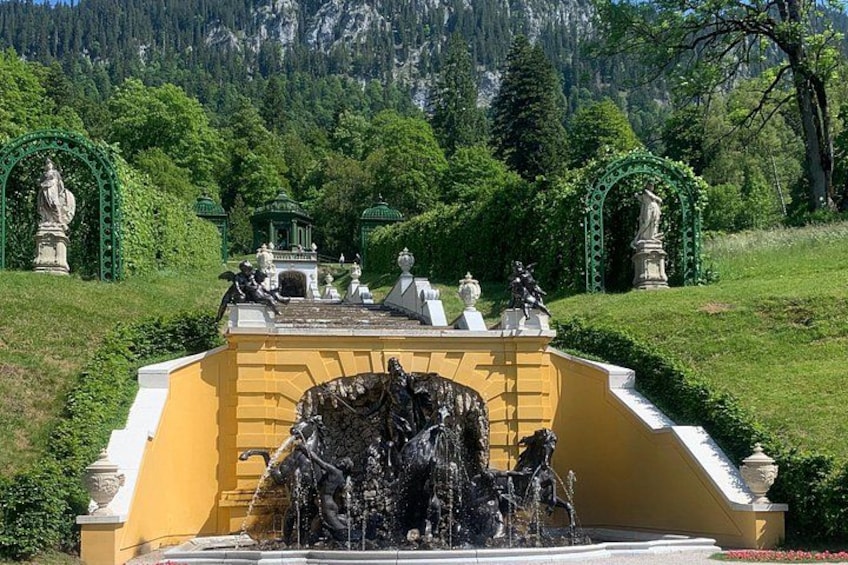 Una fuente con estatuas, un edificio amarillo y arcos verdes en un parque con árboles y montañas al fondo. 3