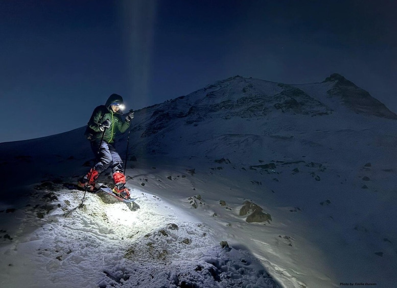 A person on skis ascending a snowy mountain slope at night with a headlamp. 1