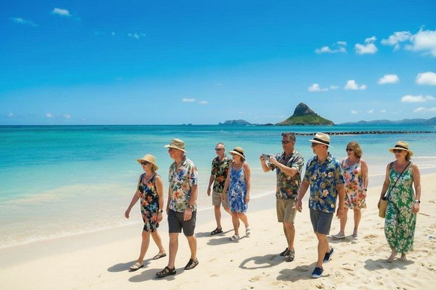 A group of people walking on a sandy beach with clear blue water and a small island in the background. 1
