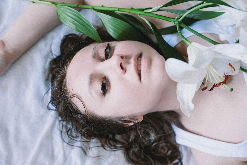 A person lying down with curly hair, wearing a white top, and holding a white flower. 5