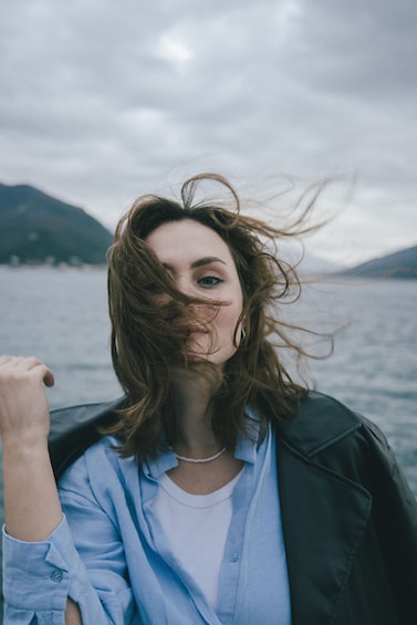 A person with curly hair, wearing a light blue shirt and a dark jacket, standing by a body of water with mountains in the background. 3