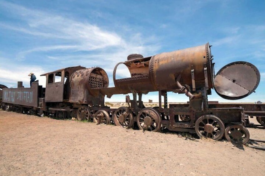 An old, rusted train engine with a large cylindrical boiler and a large wheel, situated in a barren landscape. 3