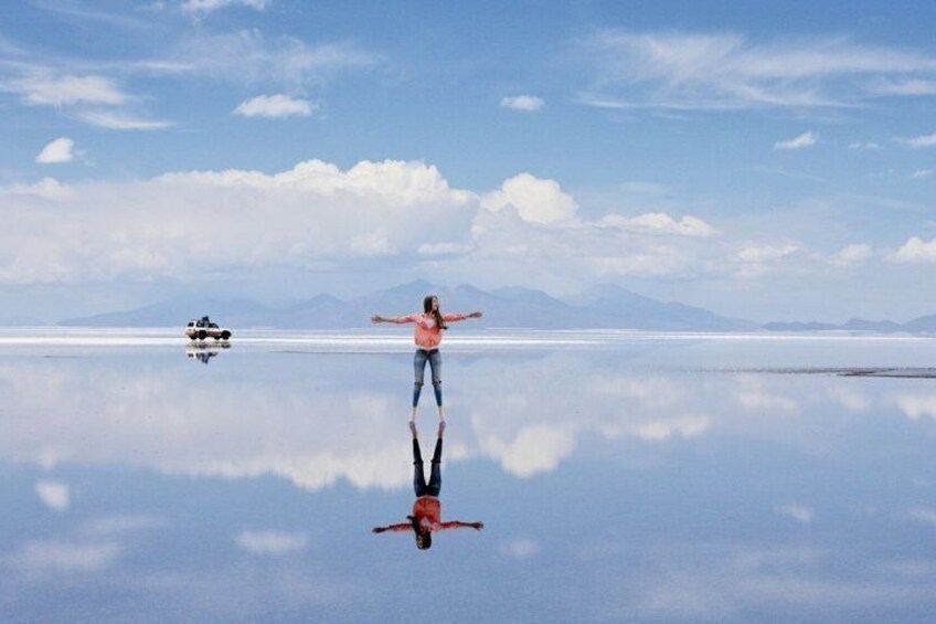 A person standing on a reflective salt flat with arms outstretched, a vehicle in the background, and mountains in the distance. 2