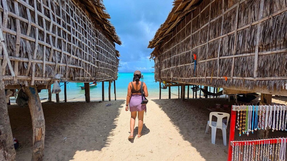 A woman walking on a sandy path between thatched-roof structures with a beach and turquoise water in the background. 1