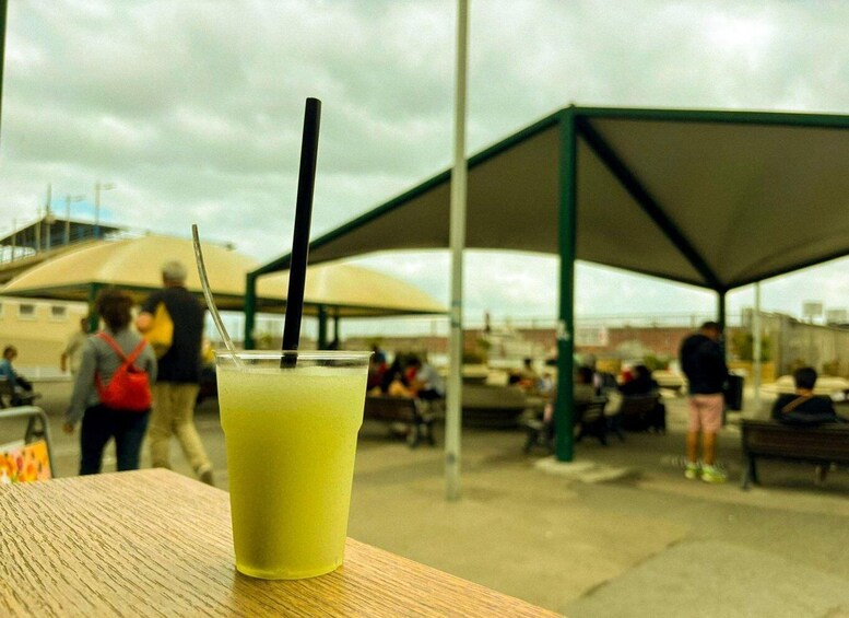 A glass of yellow beverage with a straw on a wooden table, with people and umbrellas in the background. 2