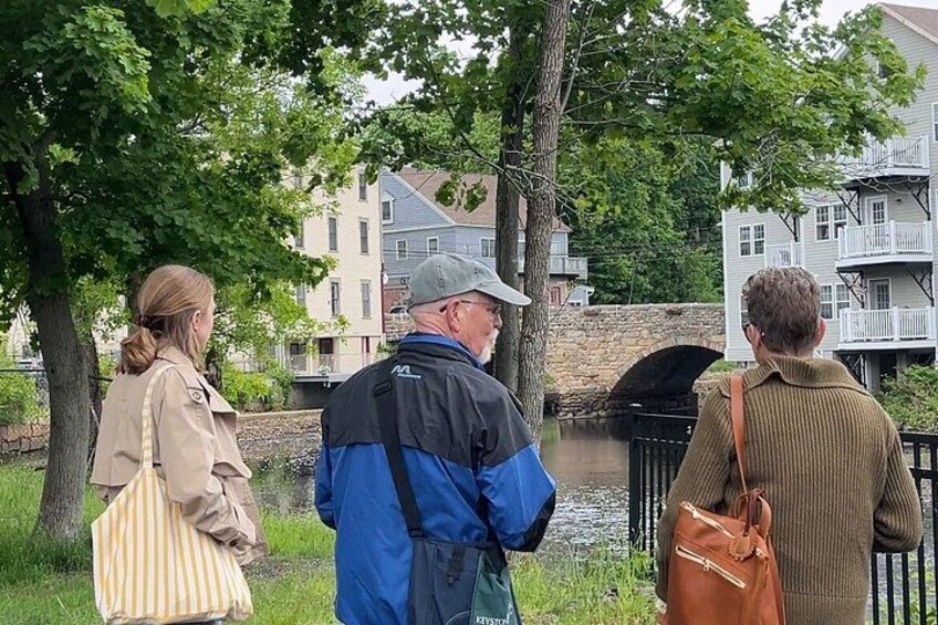 Three people standing by a canal with buildings and a stone bridge in the background. 3