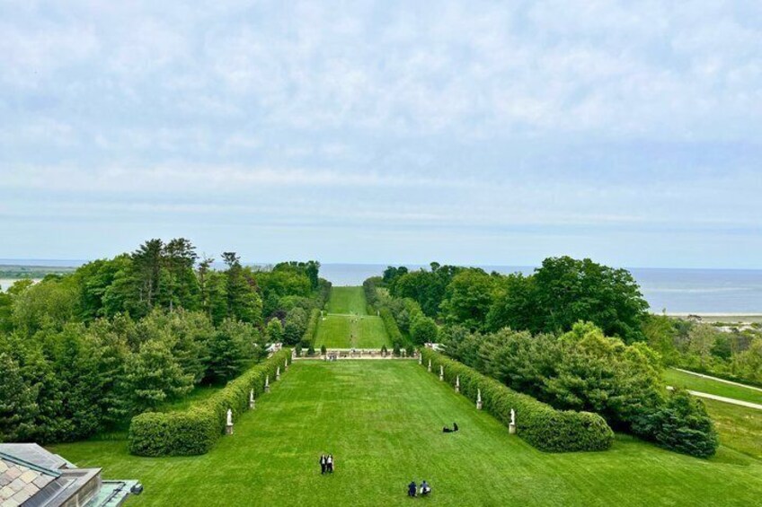 A well-manicured lawn with a hedge-lined pathway leading to a building, surrounded by trees and a view of the sea. 3