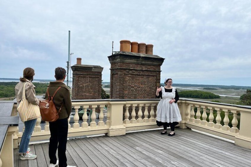 A wooden deck with a railing overlooking a landscape with brick chimneys and a distant view of the sea. 1