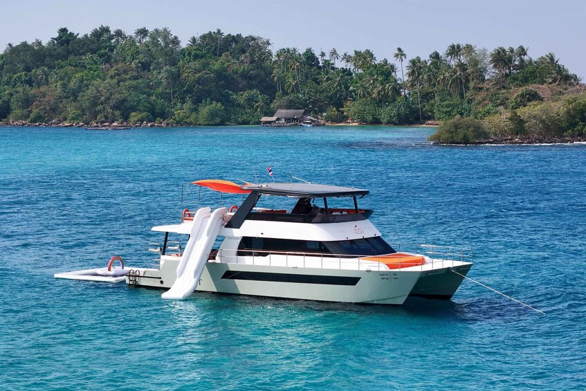 A white yacht with an orange canopy anchored in clear blue waters, surrounded by lush greenery and palm trees. 1