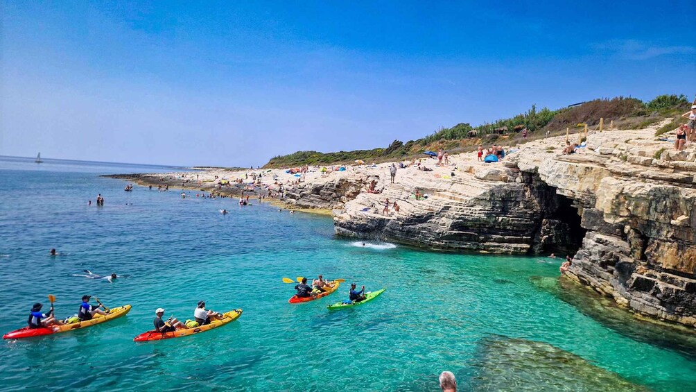 Kayakers on a clear turquoise sea, rocky cliffs, and a sandy beach with people sunbathing. 5