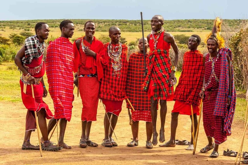 A group of people in traditional red and black patterned attire, standing on a dirt path with a grassy field and trees in the background. 3