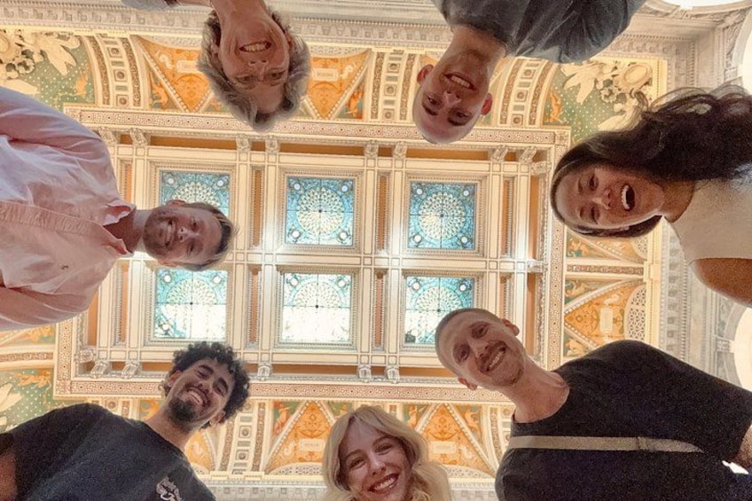 A group of people standing in a circular formation, facing a large, ornate ceiling with intricate patterns and a central stained glass window. 3