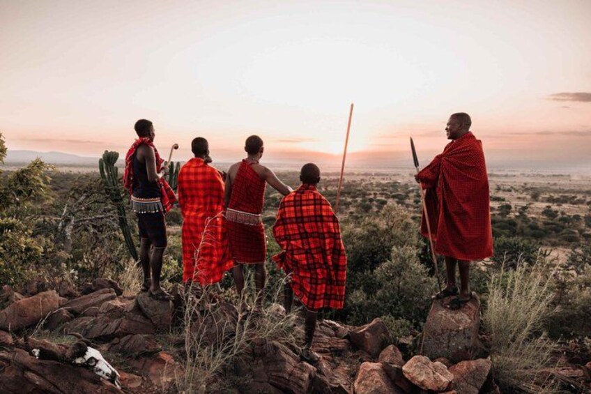 Een groep mensen in traditionele kledij staat op rotsachtig terrein en kijkt uit over een landschap met schaarse begroeiing en heuvels in de verte. 3