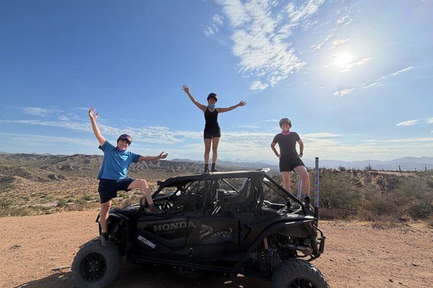 Three people on a black off-road vehicle in a desert landscape with mountains in the background. 4