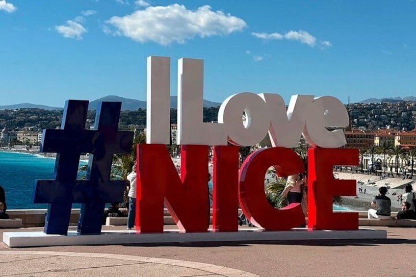 Un grande cartello esterno con la scritta 'I Love Nice' in lettere rosse e bianche, con un simbolo di hashtag e un simbolo di croce ai lati. 5
