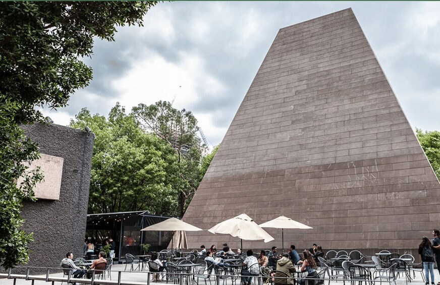 A modern building with a large triangular facade, outdoor seating with umbrellas, and trees in the background. 4