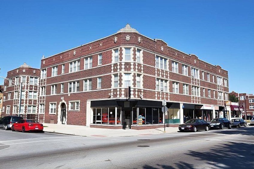 A multi-story brick building with a corner entrance, storefronts, and parked cars on a clear day. 3