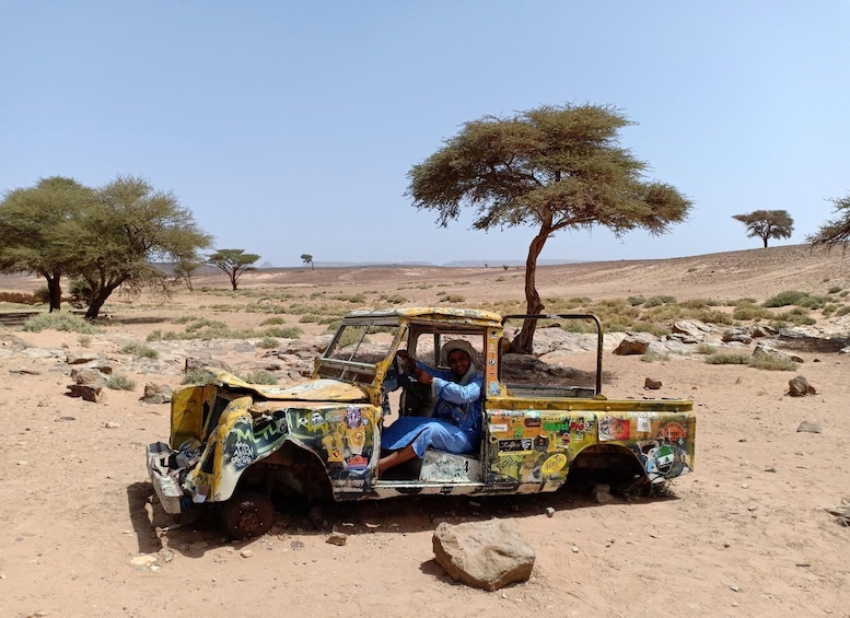 A yellow vehicle with stickers parked in a desert with sparse vegetation and a few trees. 1