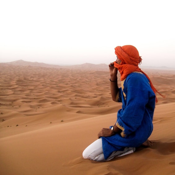 A person sitting on sand dunes, wearing a blue robe and orange headscarf, in a desert landscape. 3