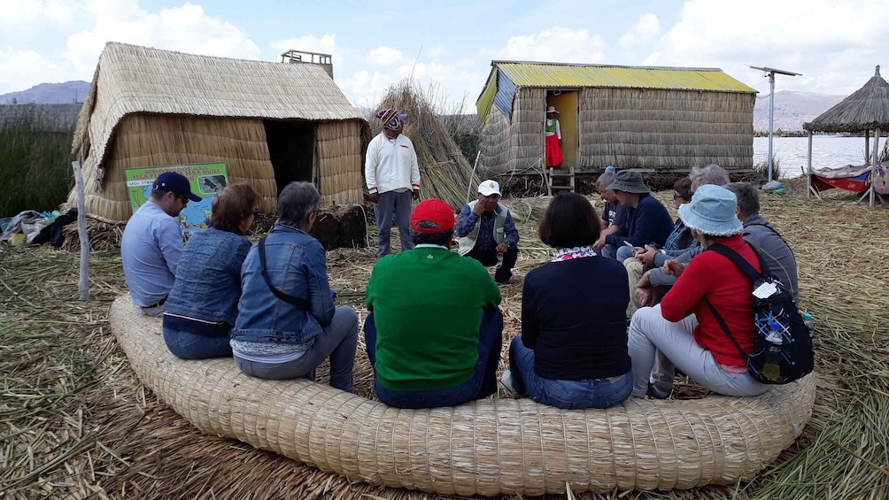 A group of people seated on a circular woven structure outdoors, with thatched-roof buildings in the background. 4
