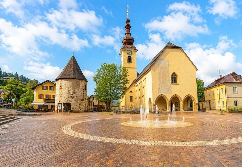 A historic square with a central fountain, a church with a clock tower, and surrounding buildings. 3