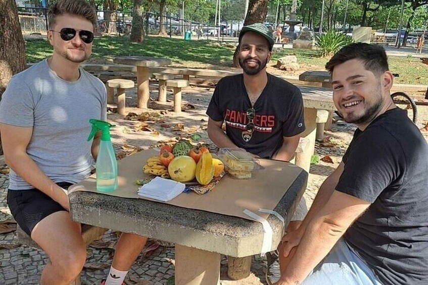 Three people sitting at a table with fruits and a spray bottle in a park. 3