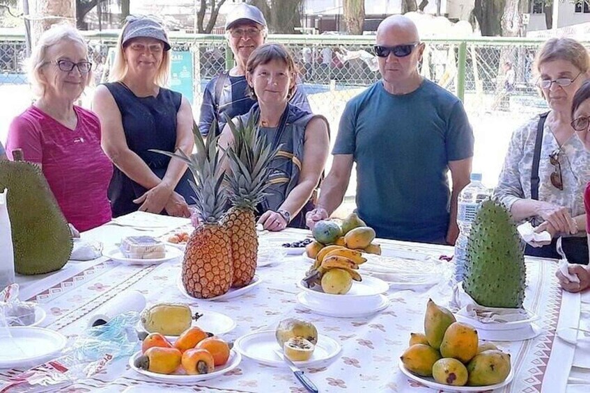 A group of people at an outdoor event with a table full of fruits like pineapples, bananas, and citrus fruits. 4