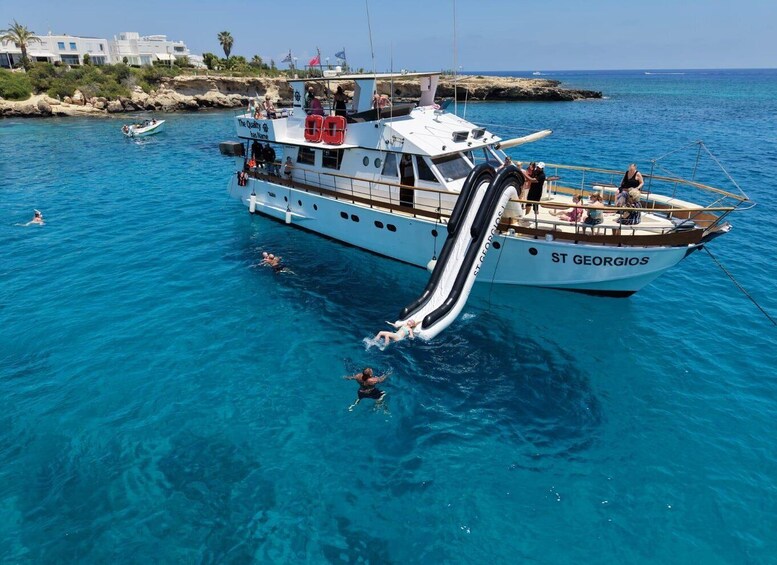 A white boat named ST GEORGIOS with a water slide, people swimming, and a coastal area with buildings in the background. 3