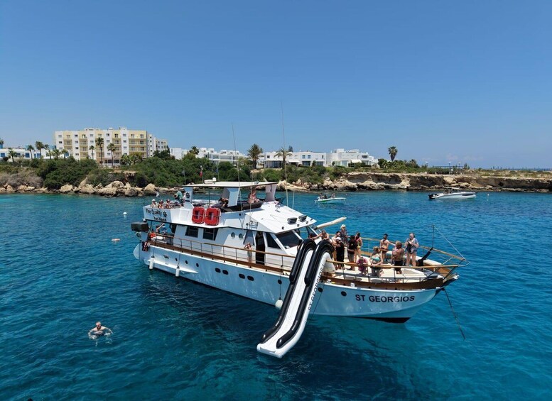 A white boat named 'St Georgios' with a slide, docked in clear blue water with people on board and others swimming nearby. 2