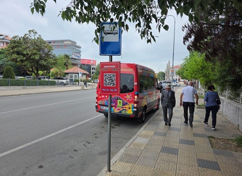 A red bus with QR code and 'SIGHTSEEING SHKODRA' on its back, parked on a city street with pedestrians walking by. 4