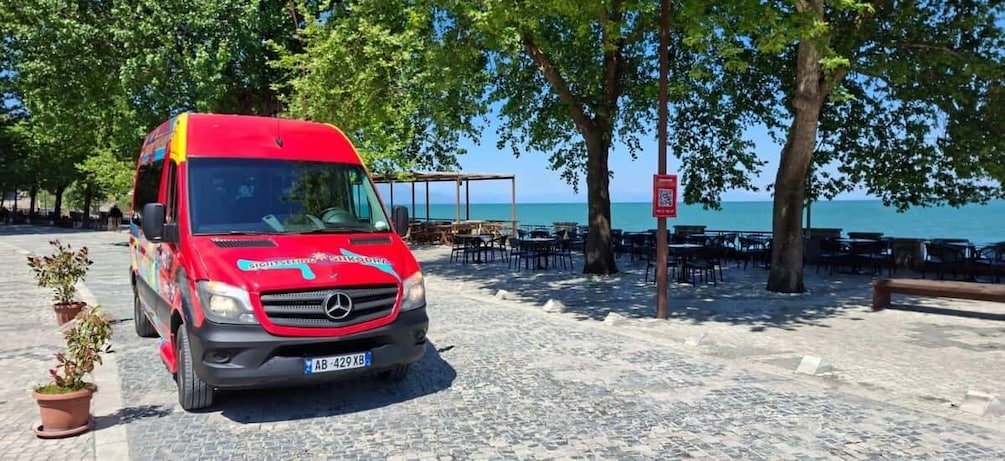 A red van parked on a cobblestone path with a beach and dining area in the background. 5