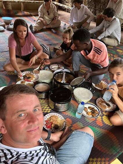 A group of people sitting on a colorful mat, eating from bowls and plates, with cooking pots and a water bottle visible. 5