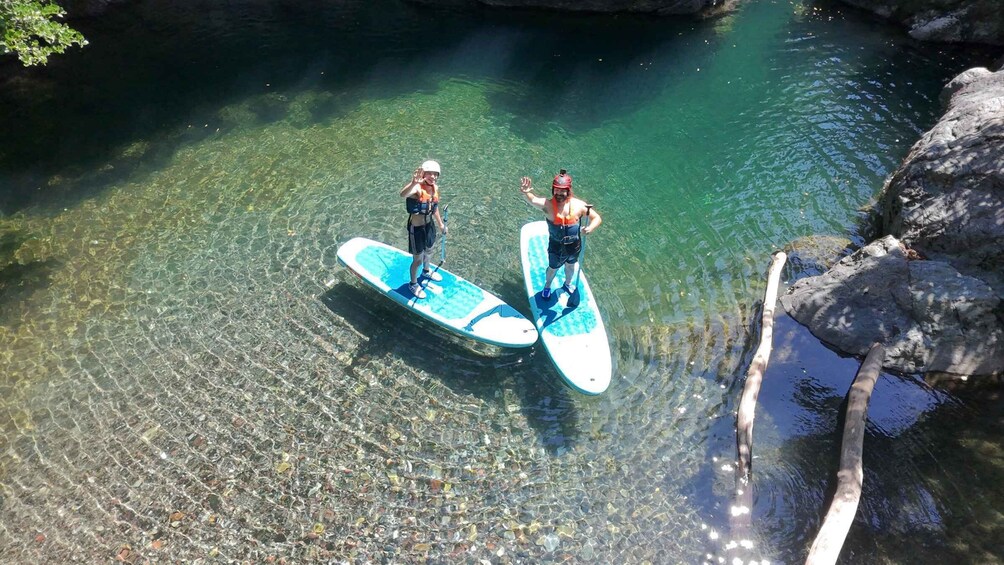 Two people paddleboarding on a clear river with rocks and trees around. 2