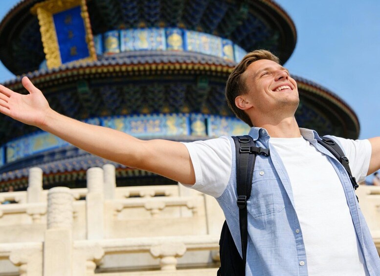 A person with arms outstretched in front of a traditional Chinese building with intricate blue and gold roof tiles. 5