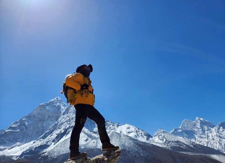 A person in a yellow jacket and black pants standing on a rocky outcrop with a mountainous landscape in the background. 5