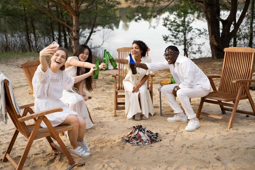 A group of people sitting on wooden chairs around a campfire on a sandy beach with trees in the background. 5