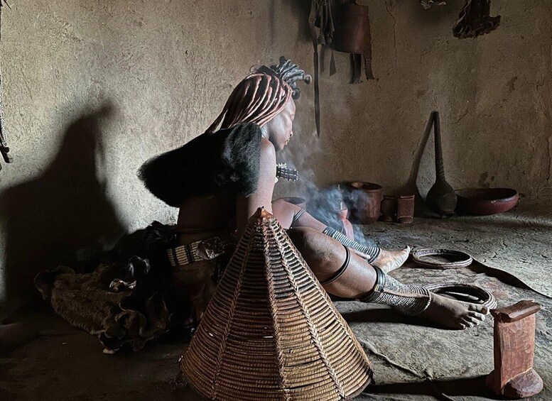 A person in traditional attire sitting on the floor with a woven basket, surrounded by pottery and a clay pot emitting steam. 1