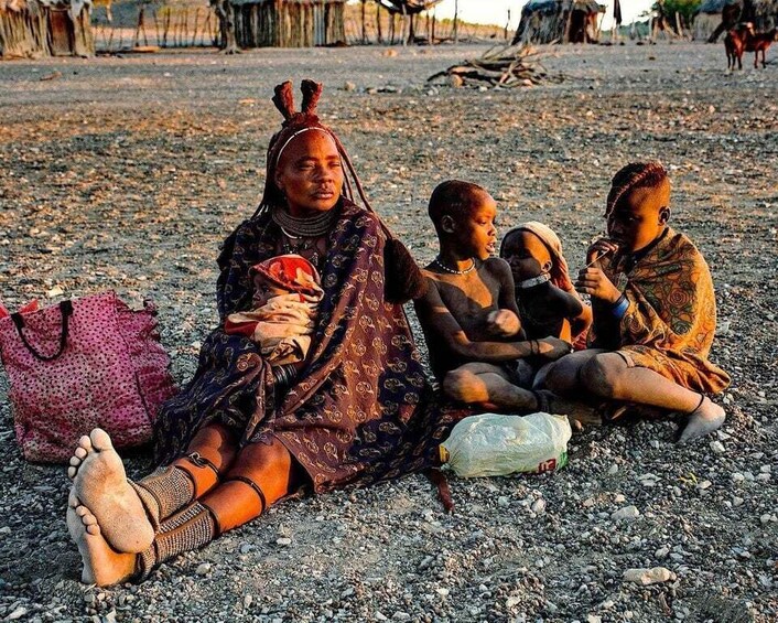 A woman and children sitting on a gravel surface, with a plastic bag and a pink bag nearby. 2