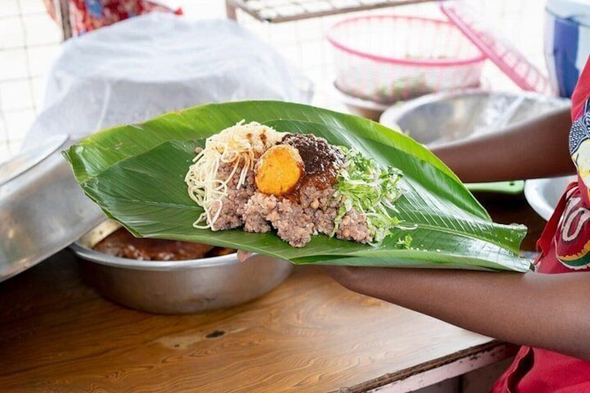 A person holding a food dish on a banana leaf, with a metal bowl and a plastic container in the background. 4